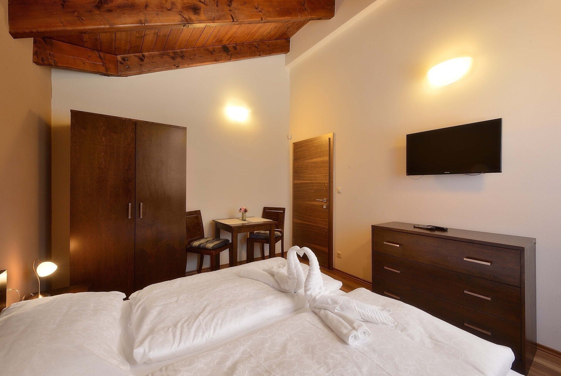 Une chambre d’hôtel avec un plafond en bois, un lit recouvert de draps blancs, une commode en bois, un téléviseur fixé au mur et un coin repas avec une table et des chaises.