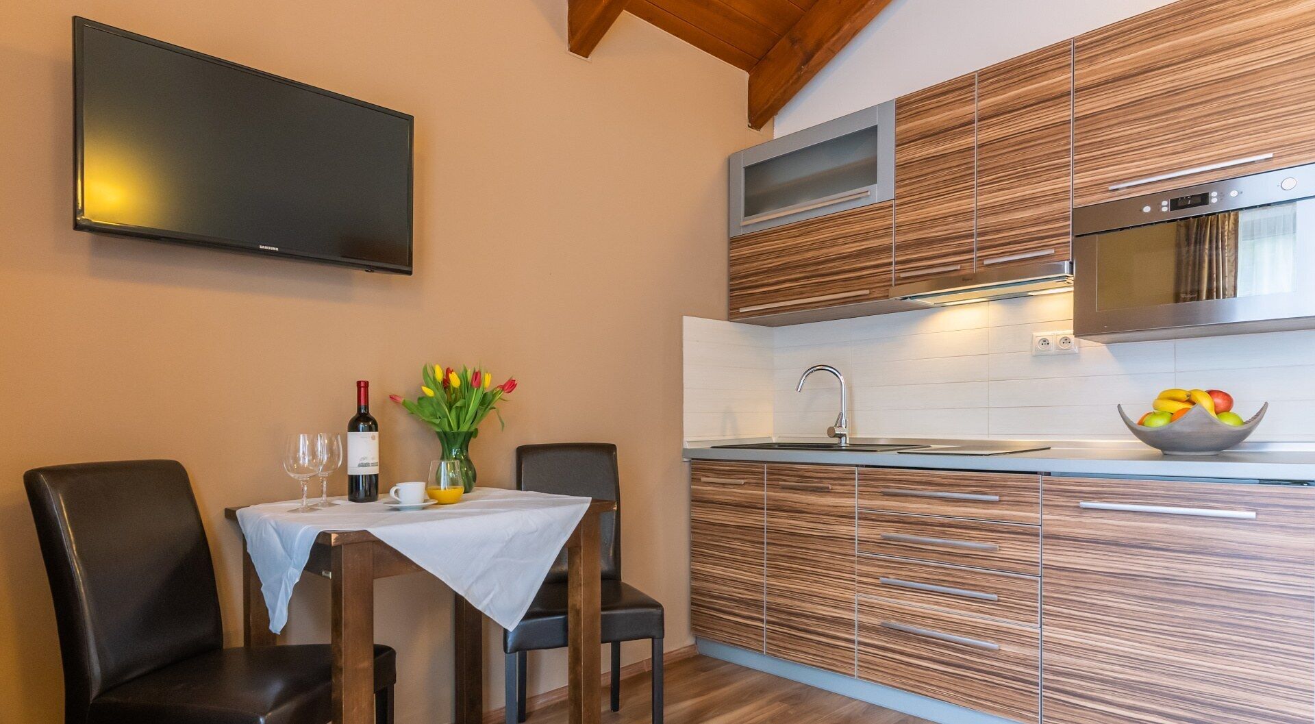 A modern kitchen with wooden cabinetry, a built-in microwave, and a large flat-screen TV mounted on the wall.