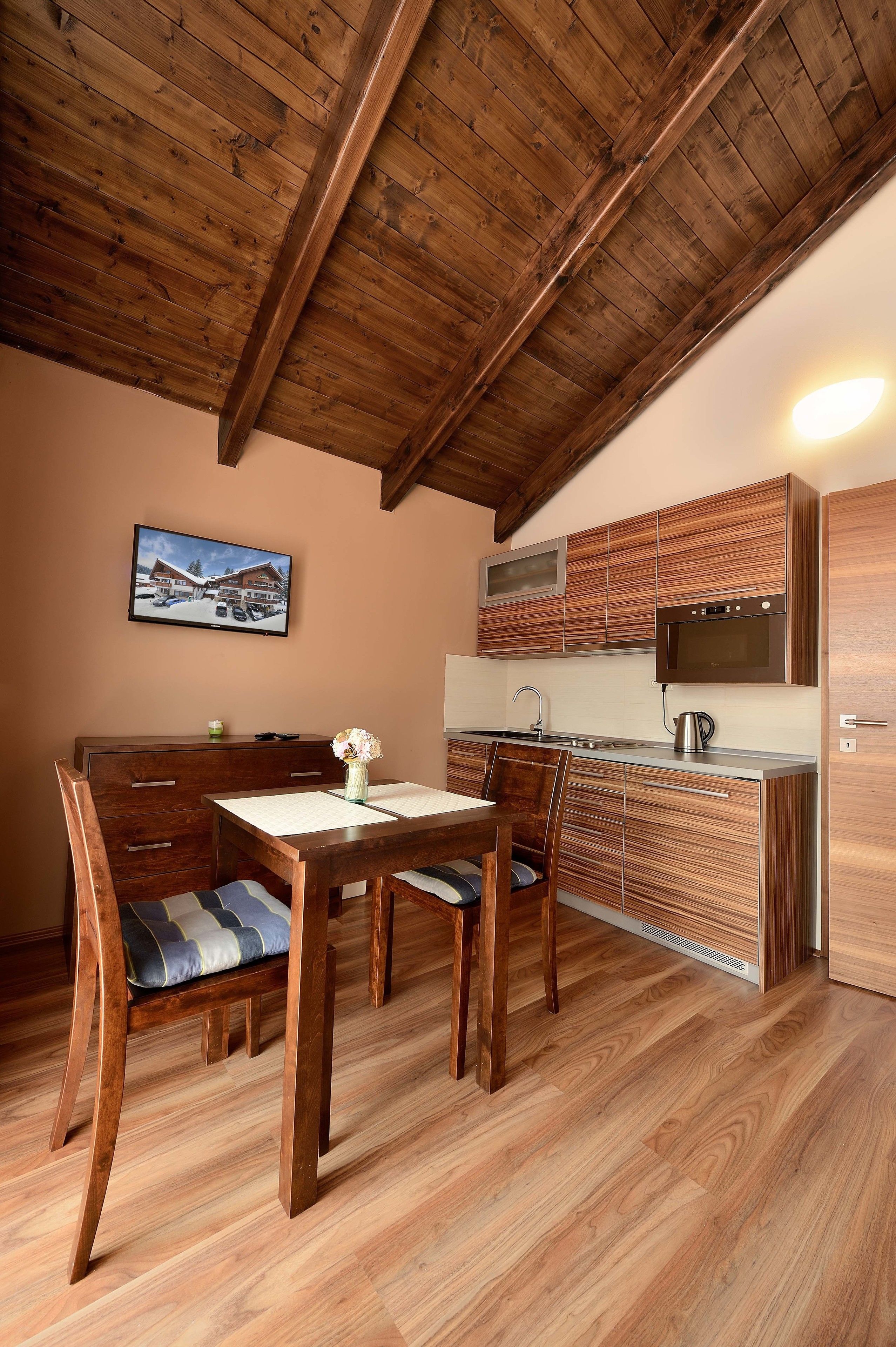 Una mesa de comedor de madera con sillas, una cocina con armarios de madera y un televisor en la pared.