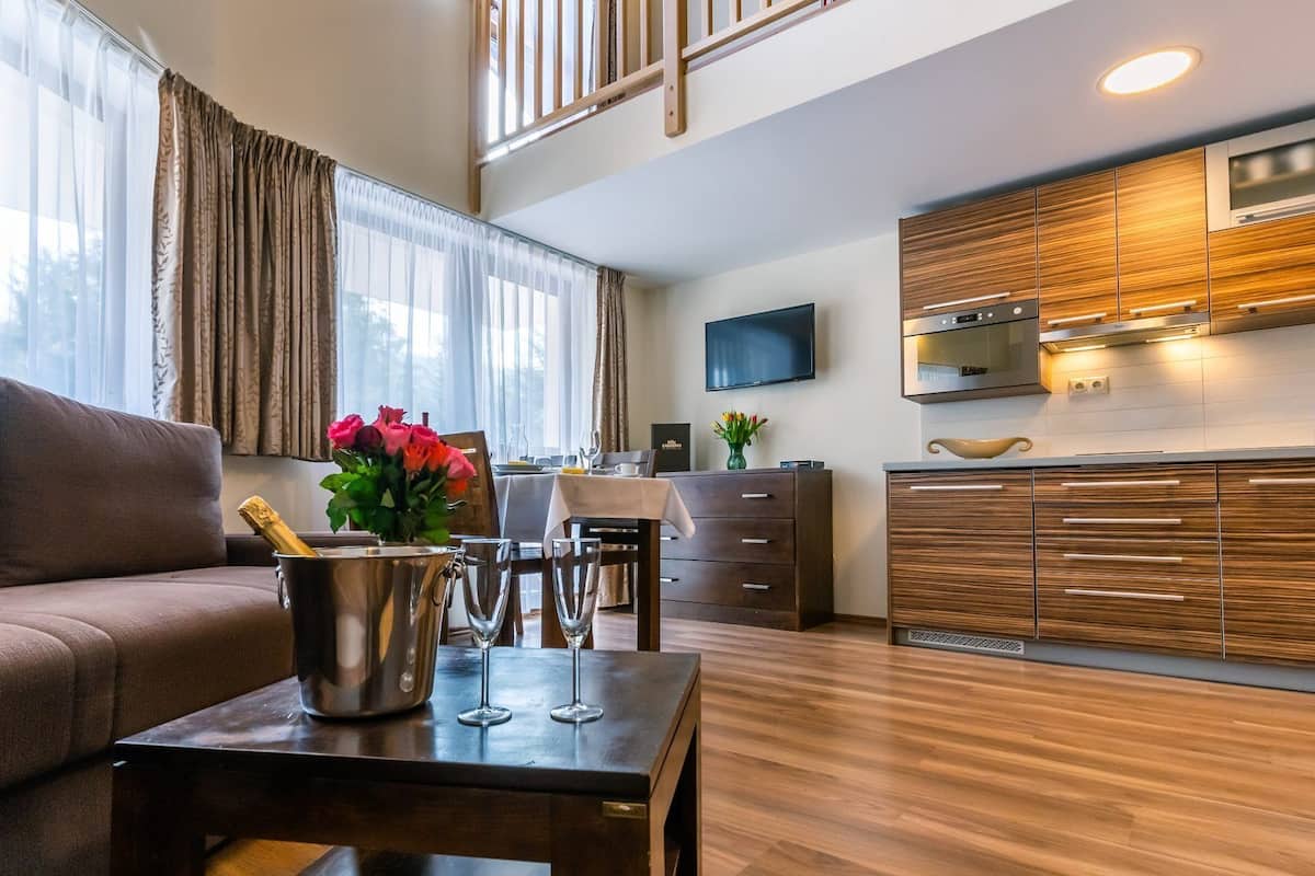 A modern living room with a sofa, a dining table with a champagne bottle and glasses, and a kitchen with wooden cabinets and a TV.