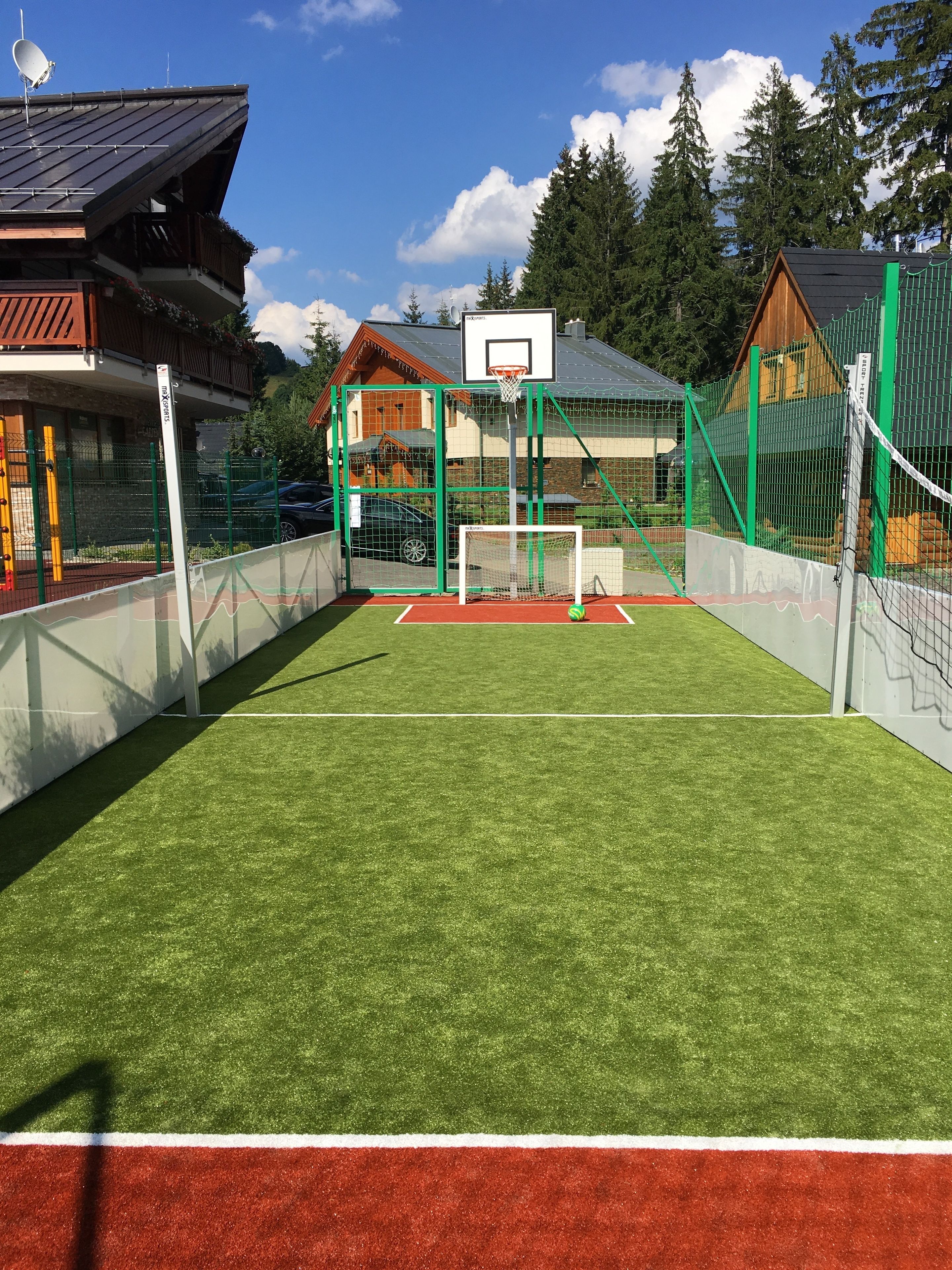 Een buitensportveld met een basketbalring, omringd door groen hekwerk en bomen.