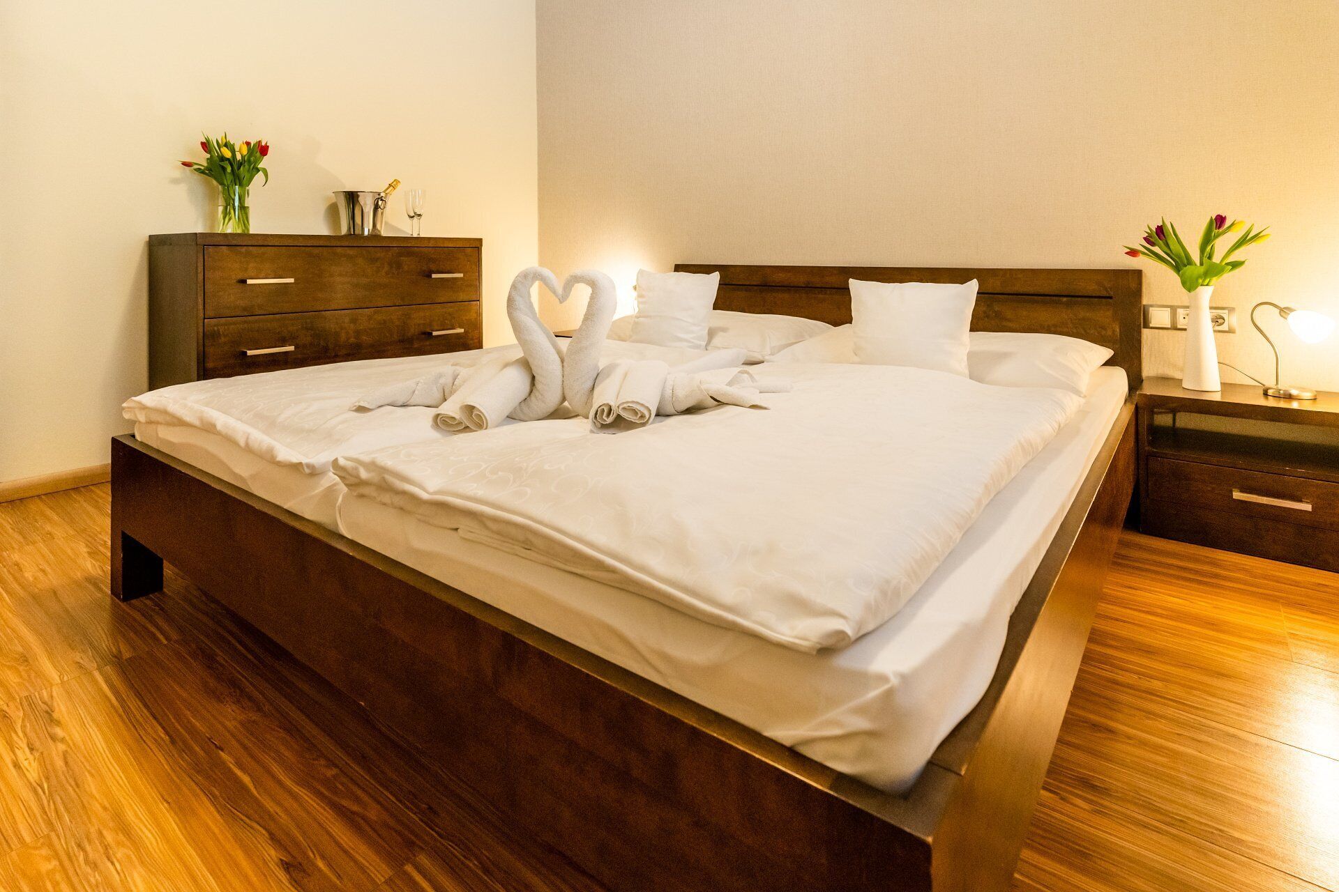 A neatly made bed with towels arranged in a heart shape, a wooden nightstand with a lamp, and a vase with flowers on the nightstand.