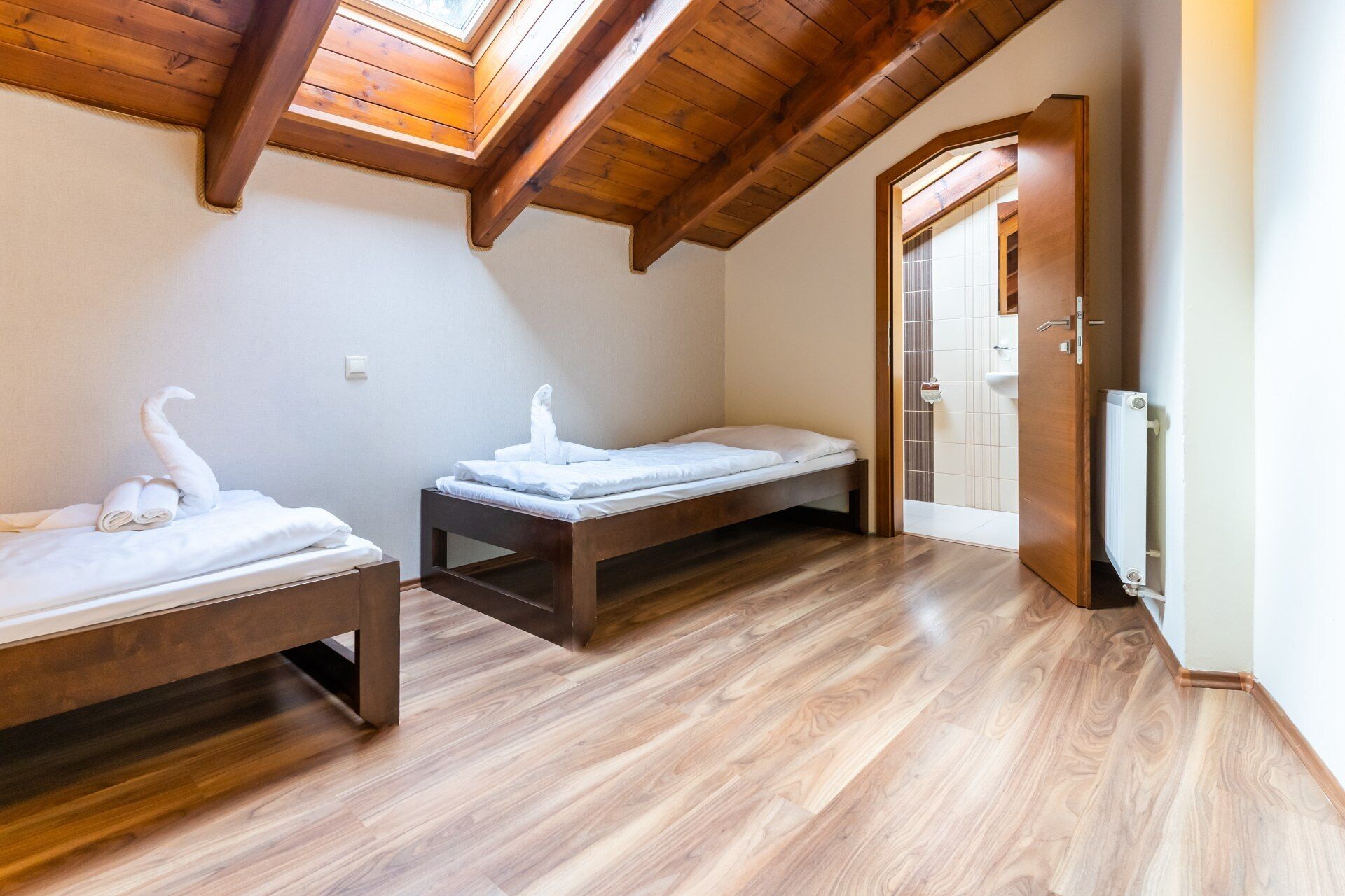 A room with two beds, wooden flooring, a skylight, and a door leading to a bathroom.
