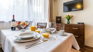 Habitación de hotel con preparación para el desayuno, incluyendo una botella de vino, frutero, zumo de naranja y una mesa de comedor con platos, cubiertos y vasos.
