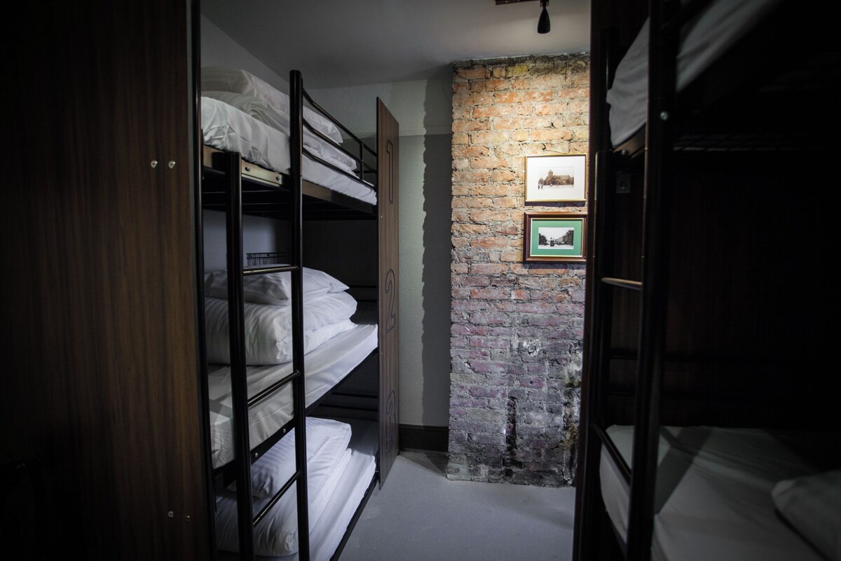A narrow hallway with bunk beds on either side, a brick wall with framed pictures, and a wooden door.