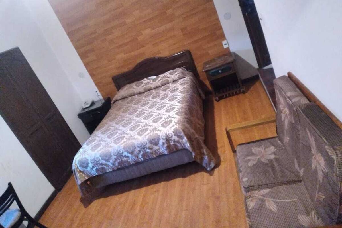 Un dormitorio con piso de madera, una cama con cubrecama estampado, una mesita de noche pequeña y una silla con cojín floral.
