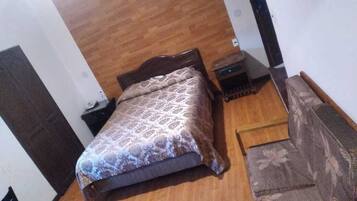 A bedroom with a wooden floor, a bed with a patterned bedspread, a small nightstand, and a chair with a floral cushion.