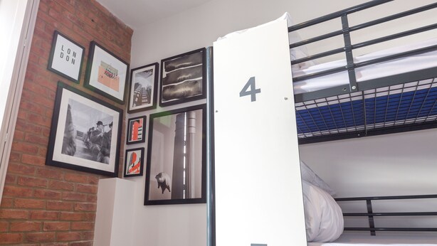 A dormitory room with bunk beds, numbered 4 and 5, and a brick wall with framed pictures. - PubLove @ The Green Man - Hostel (London)