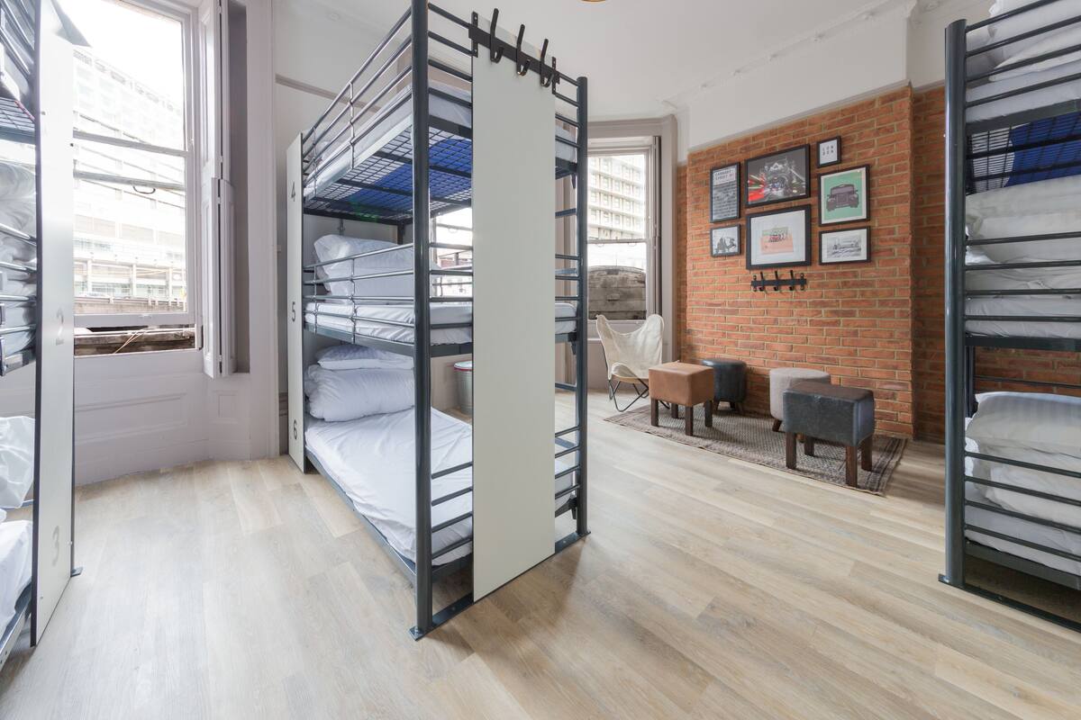 Une chambre d’hôtel avec des lits superposés, un mur de briques orné de tableaux encadrés et un petit coin salon avec un fauteuil et un pouf.