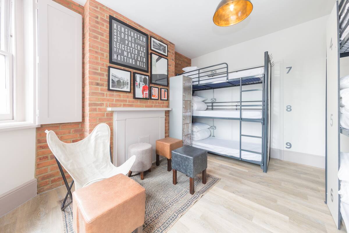 Une chambre moderne avec un lit superposé, une chaise blanche, un petit pouf, un tapis à motifs et un mur en briques orné de tableaux encadrés.