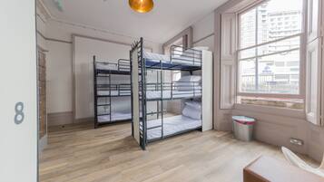 A room with bunk beds, a wooden floor, a window with a view of buildings, and a trash can.