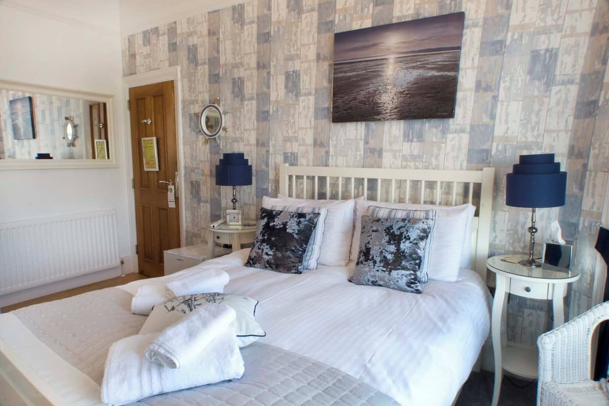 A neatly made bed with white linens and two pillows, a bedside table with a lamp, a mirror, and a framed picture on the wall.