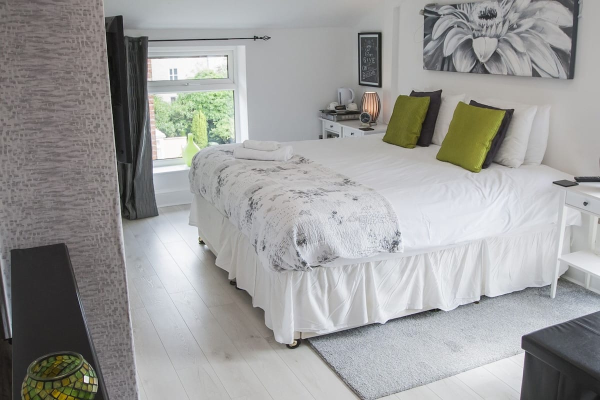 A modern bedroom with a large bed, a bedside table with a lamp, a small desk, and a window with curtains.