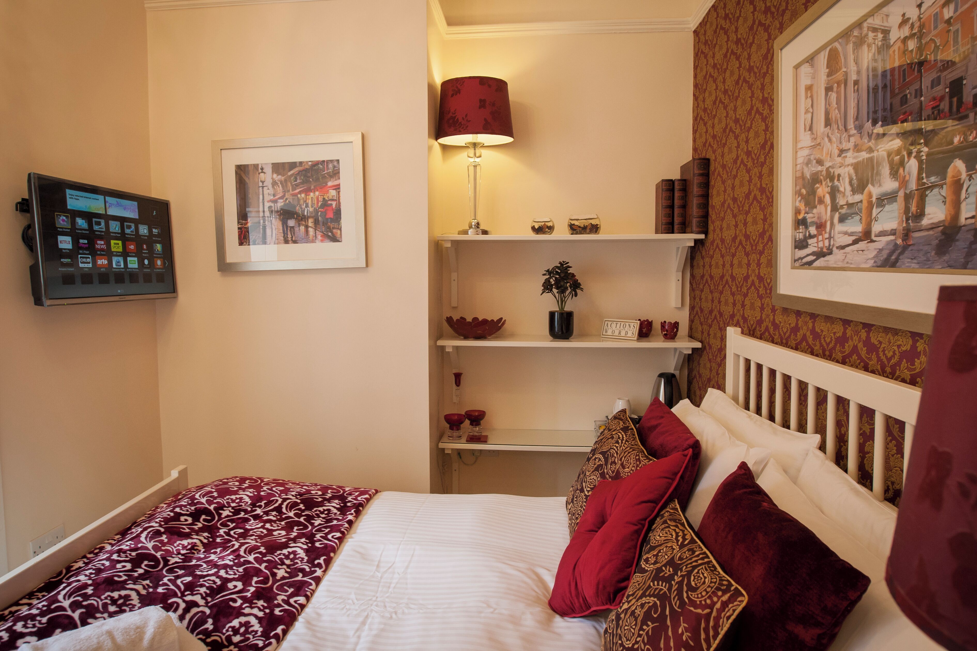 Habitación de hotel con cama, mesita de noche, lámpara, un cuadro en la pared y estante con adornos.