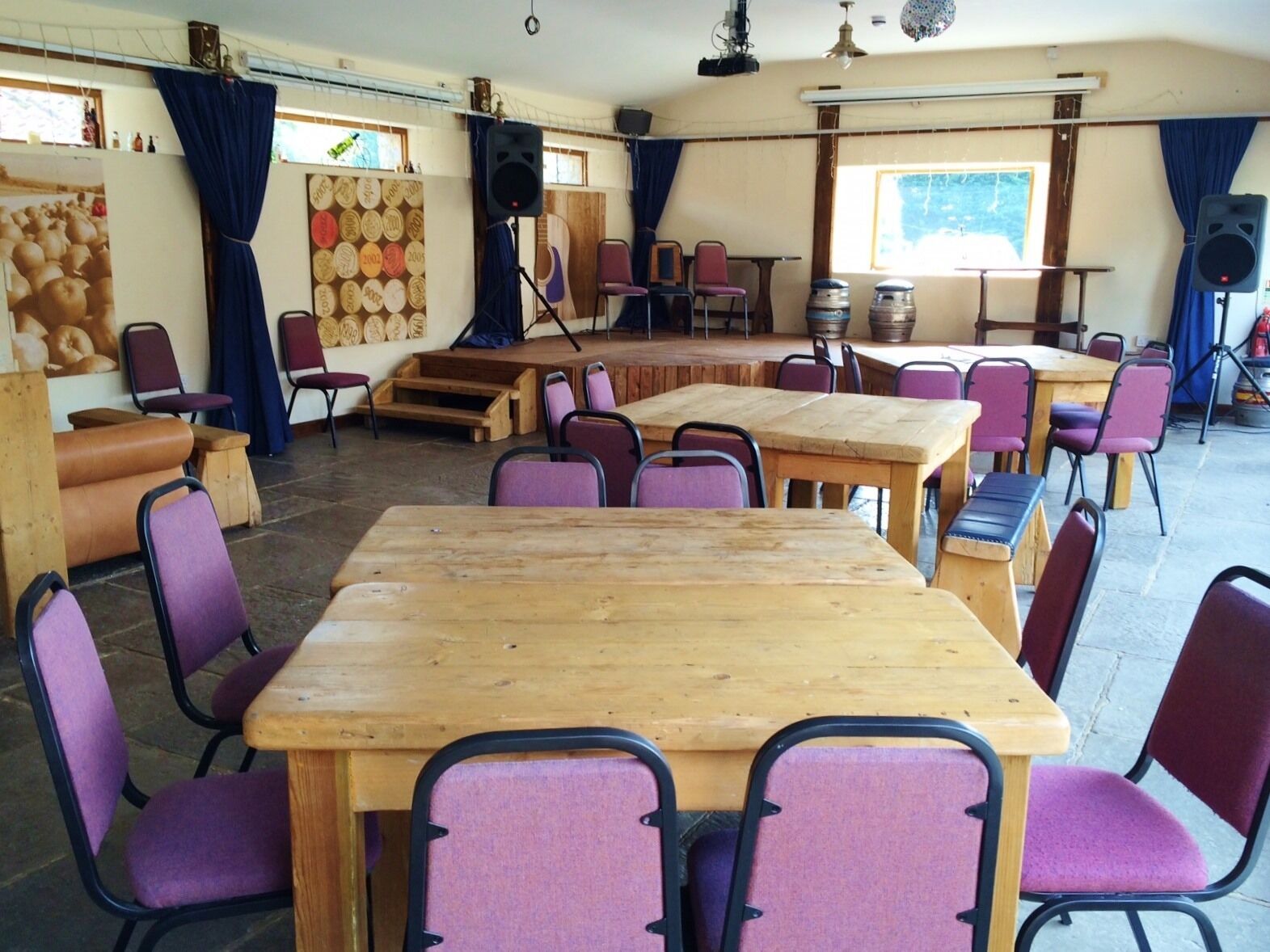A room with multiple wooden tables and purple chairs, arranged in a semi-circle. There are large speakers and a stage area with a backdrop.