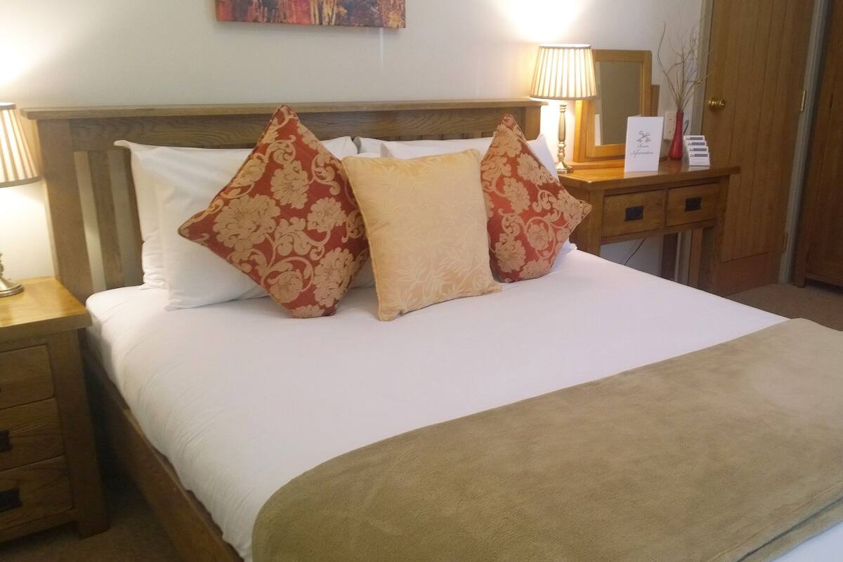 A neatly made bed with a beige blanket and three patterned pillows. There is a wooden nightstand with a lamp and a small table with a mirror and a vase with branches.