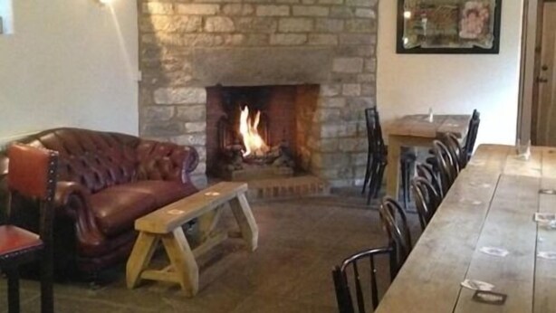 A stone fireplace with a fire, a leather sofa, and a wooden dining table with chairs. - The Cross Keys Inn (Somerton)