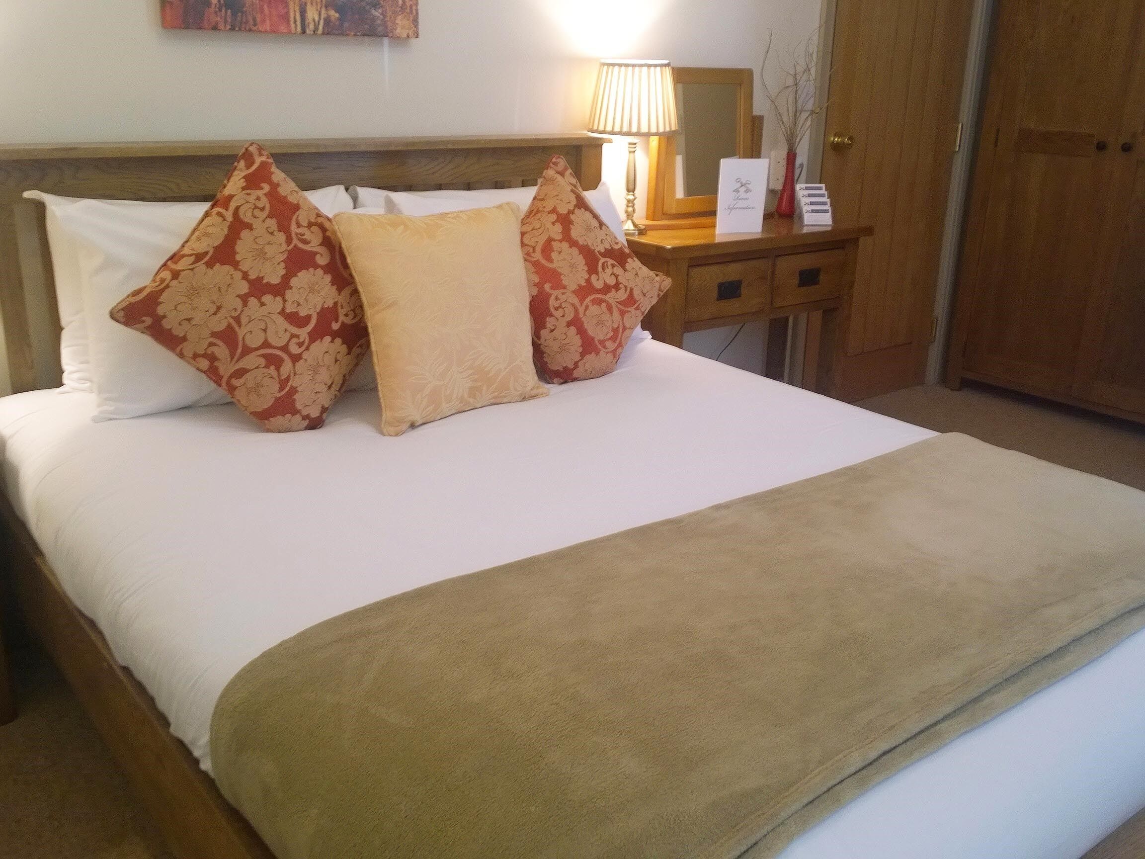 Una cama bien hecha con almohadas de estampado floral, una mesita de noche con lámpara y una puerta de madera al fondo.