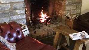 A cozy living room with a fireplace, a leather sofa, and a wooden table with a newspaper. - The Cross Keys Inn (Somerton)