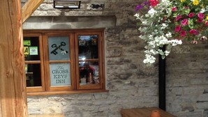 A rustic inn with a stone wall exterior, wooden tables and benches, and a window displaying a sign for 'The Cross Keys Inn'. - The Cross Keys Inn (Somerton)