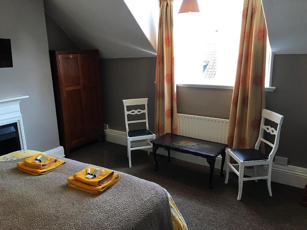 Quarto com uma cama, duas cadeiras brancas, uma mesa pequena, um guarda-roupa de madeira e uma janela com cortinas.