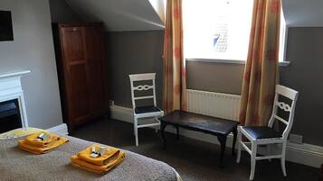 Quarto com uma cama, duas cadeiras brancas, uma mesa pequena, um guarda-roupa de madeira e uma janela com cortinas.
