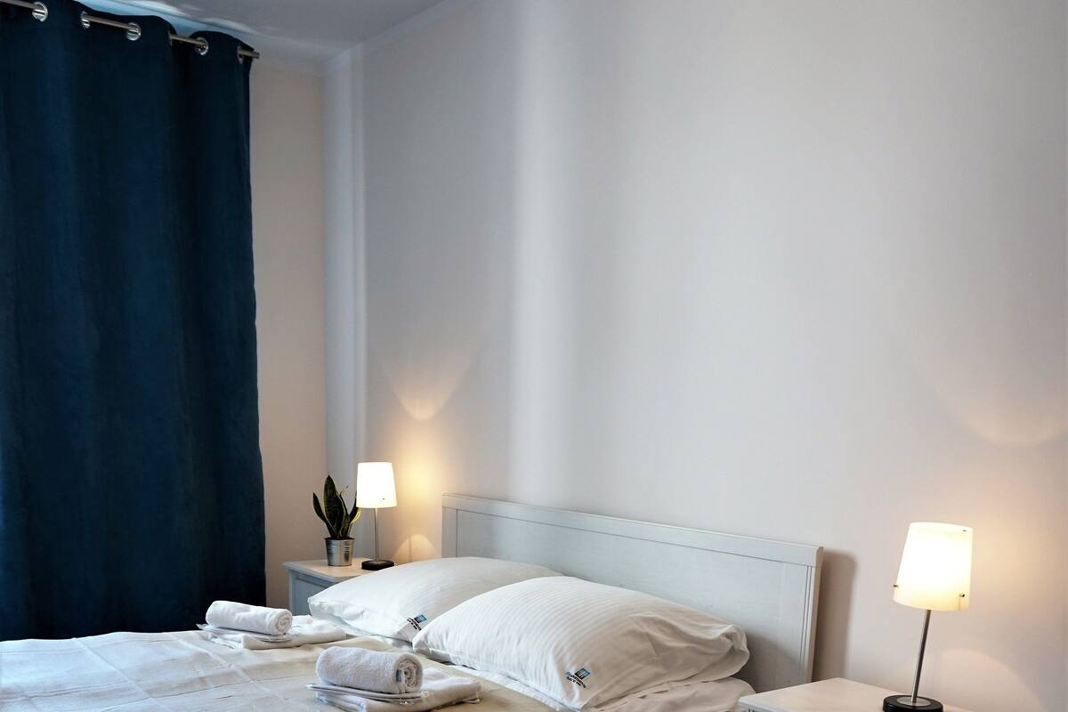 Una cama bien hecha con sábanas blancas, un tapete verde estampado y dos lámparas de noche en una mesita blanca.