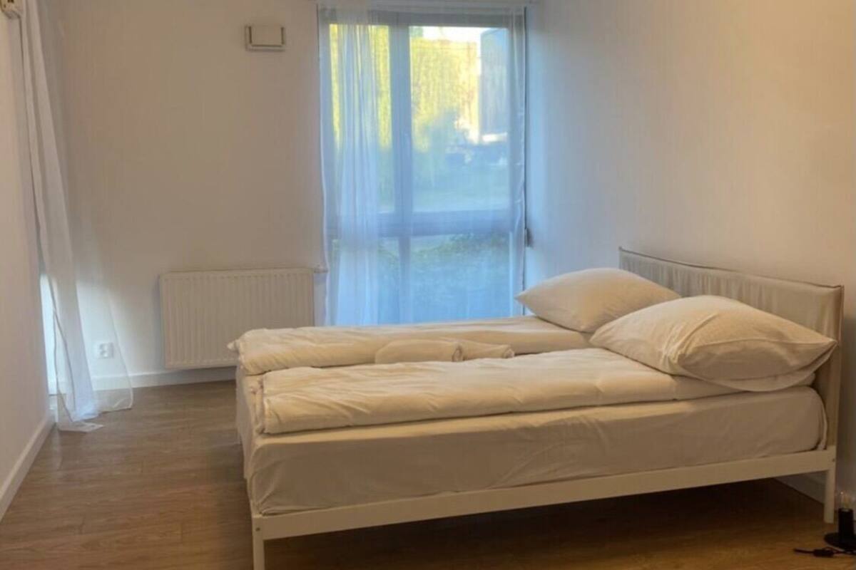 Una cama individual con ropa de cama blanca en una habitación con suelo de madera y un ventanal con cortinas de encaje.