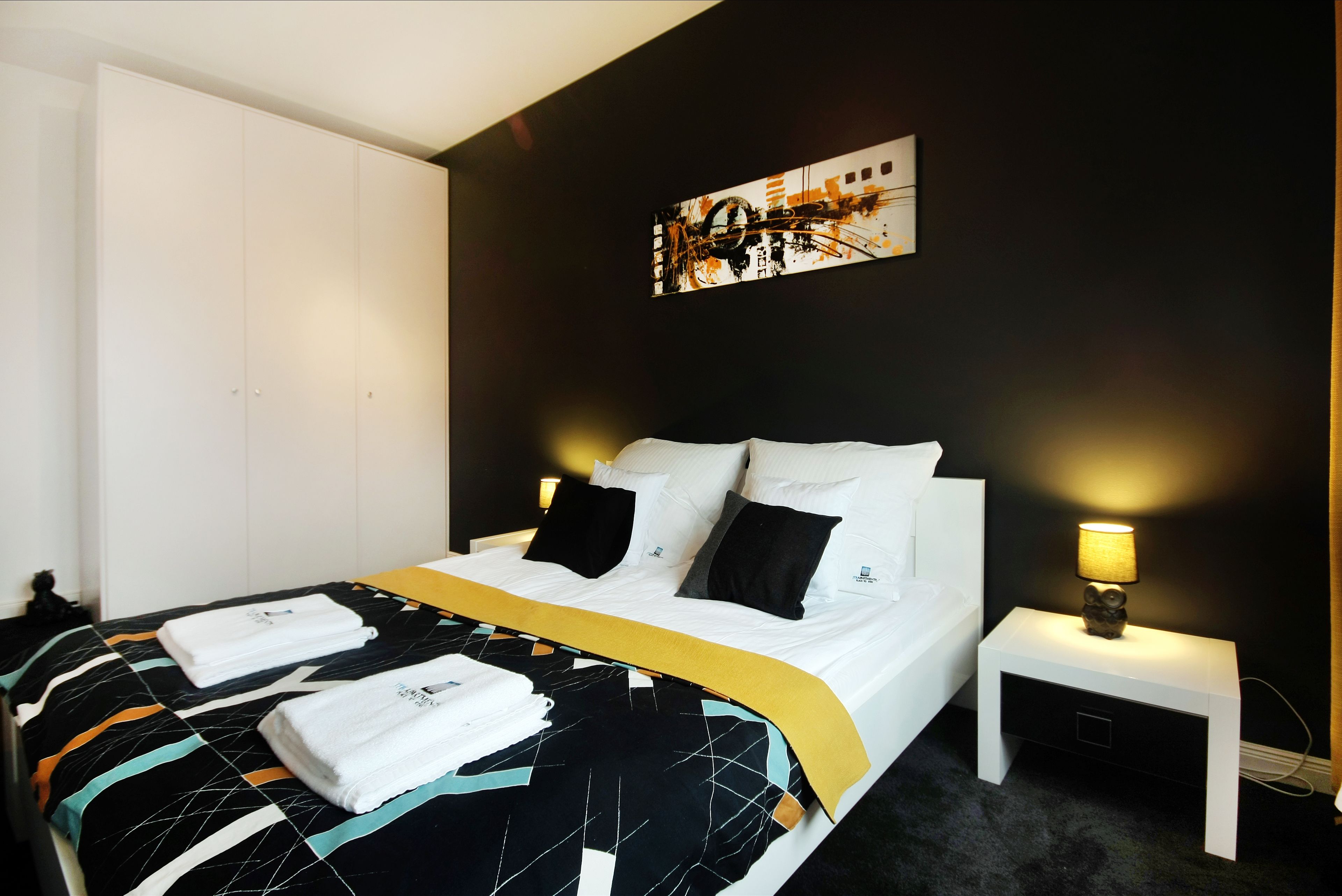 Ein ordentlich bezogenes Bett mit weißen Leinen, einer schwarz-weißen Bettdecke und einem gelben Akzent. Ein kleiner Nachttisch mit einer Lampe und ein Wandbild über dem Bett sind ebenfalls zu sehen.