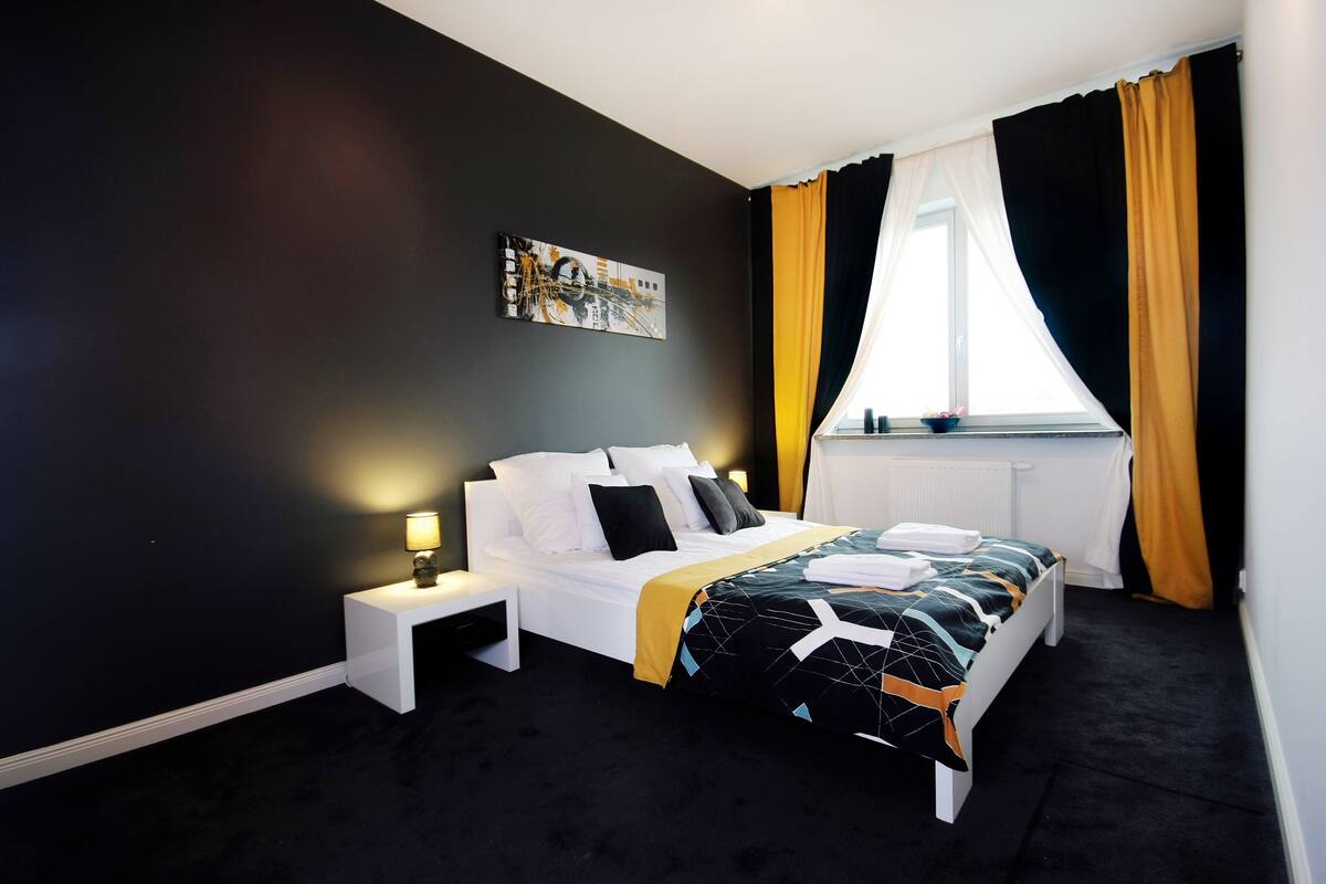 Una habitación de hotel moderna con una cama grande, una mesita de noche con lámpara, una ventana con cortinas amarillas y un cuadro en la pared.