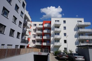 Un immeuble résidentiel à plusieurs étages, doté de balcons, avec une structure centrale de couleur rouge et un espace de stationnement où sont garées des voitures.