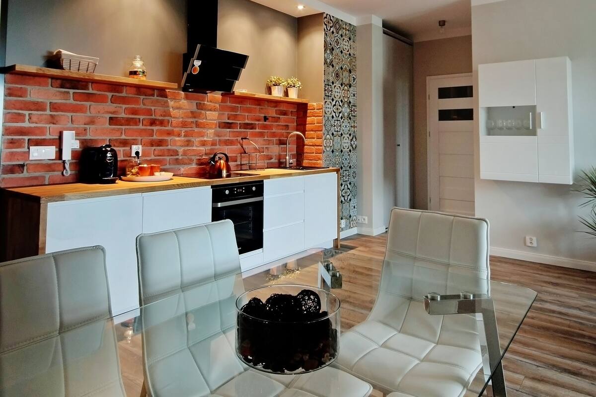 Una cocina moderna con salpicadero de ladrillo, una isla central y un área de comedor con sillas blancas y una mesa de vidrio.
