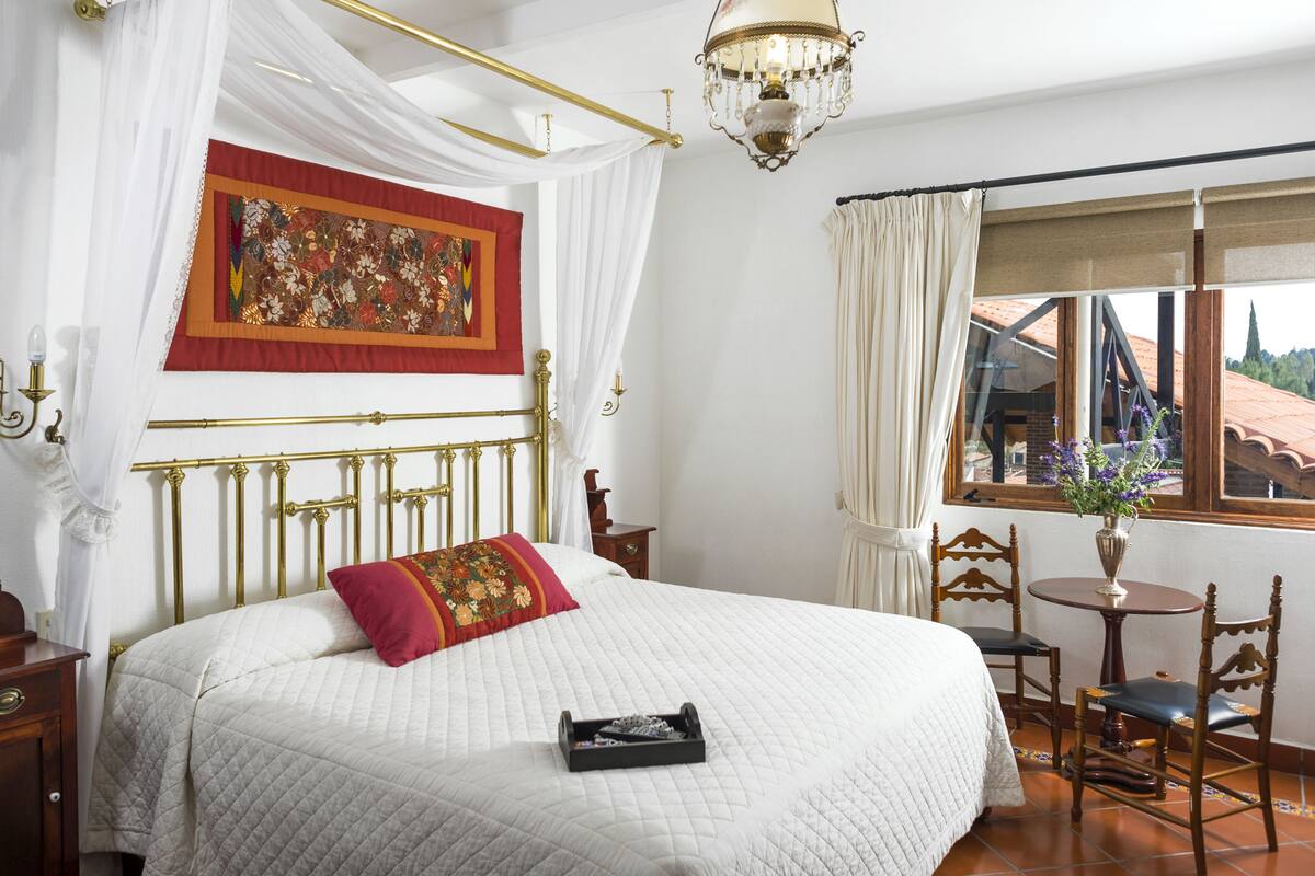 Un dormitorio con una cama con dosel, un candelabro, un ventanal con vistas y una mesa con sillas.