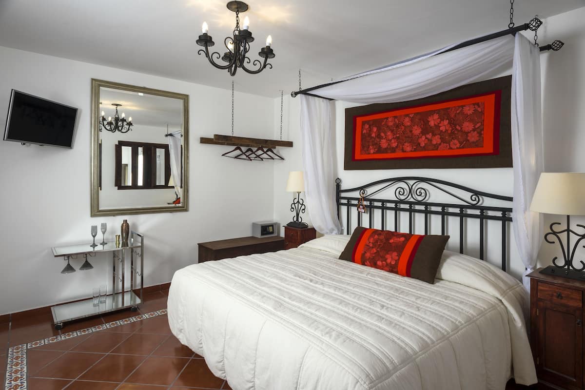 Un dormitorio ordenado con una cama grande, una mesita de noche, un velador y un televisor montado en la pared, además de una araña.