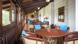 Un porche cubierto con vigas de madera, paredes de ladrillo y sillas con cojines azules alrededor de una mesa central con una planta en maceta.