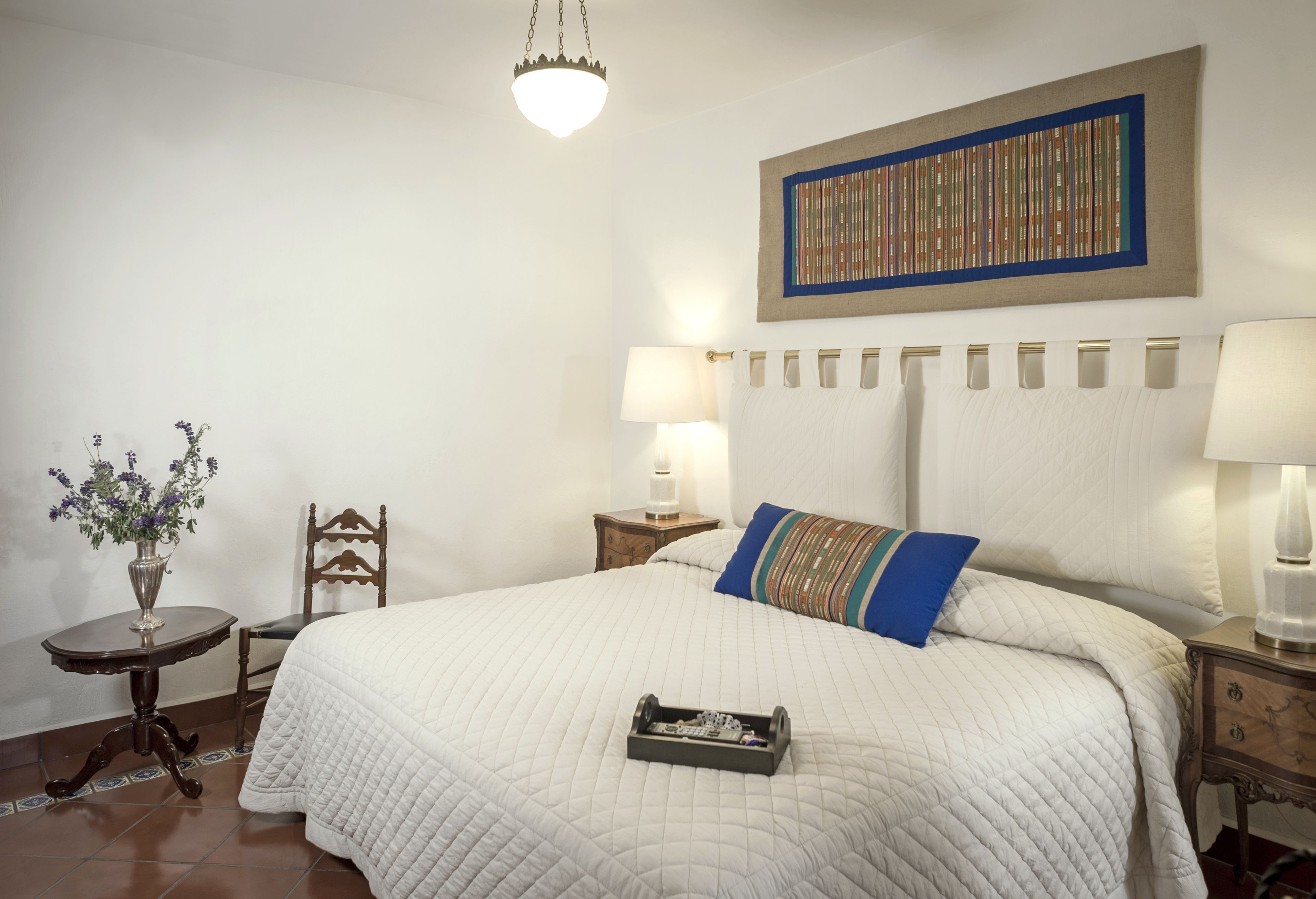 Un dormitorio ordenado con una cama grande, una mesita de noche, una silla, una lámpara y una obra de arte en la pared.