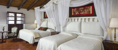 Una habitación con dos camas, cada una con ropa de cama blanca y dosel. Hay una silla de madera y una mesita con una lámpara. El suelo es de baldosas y hay un candelabro.