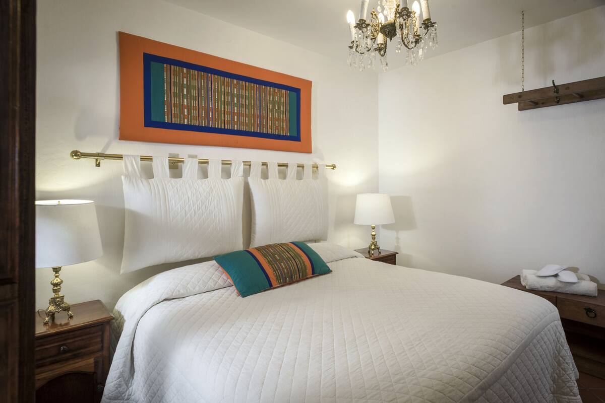 Una cama bien hecha con un edredón blanco acolchado, dos almohadas blancas y una almohada decorativa de colores. Encima de la cama hay una obra de arte con marco dorado, un candelabro colgando del techo y una estantería de madera con toallas al lado derecho.