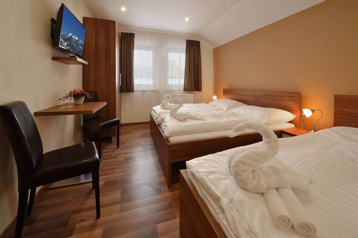 A hotel room with a wooden desk, two beds, a TV mounted on the wall, and a window with curtains.