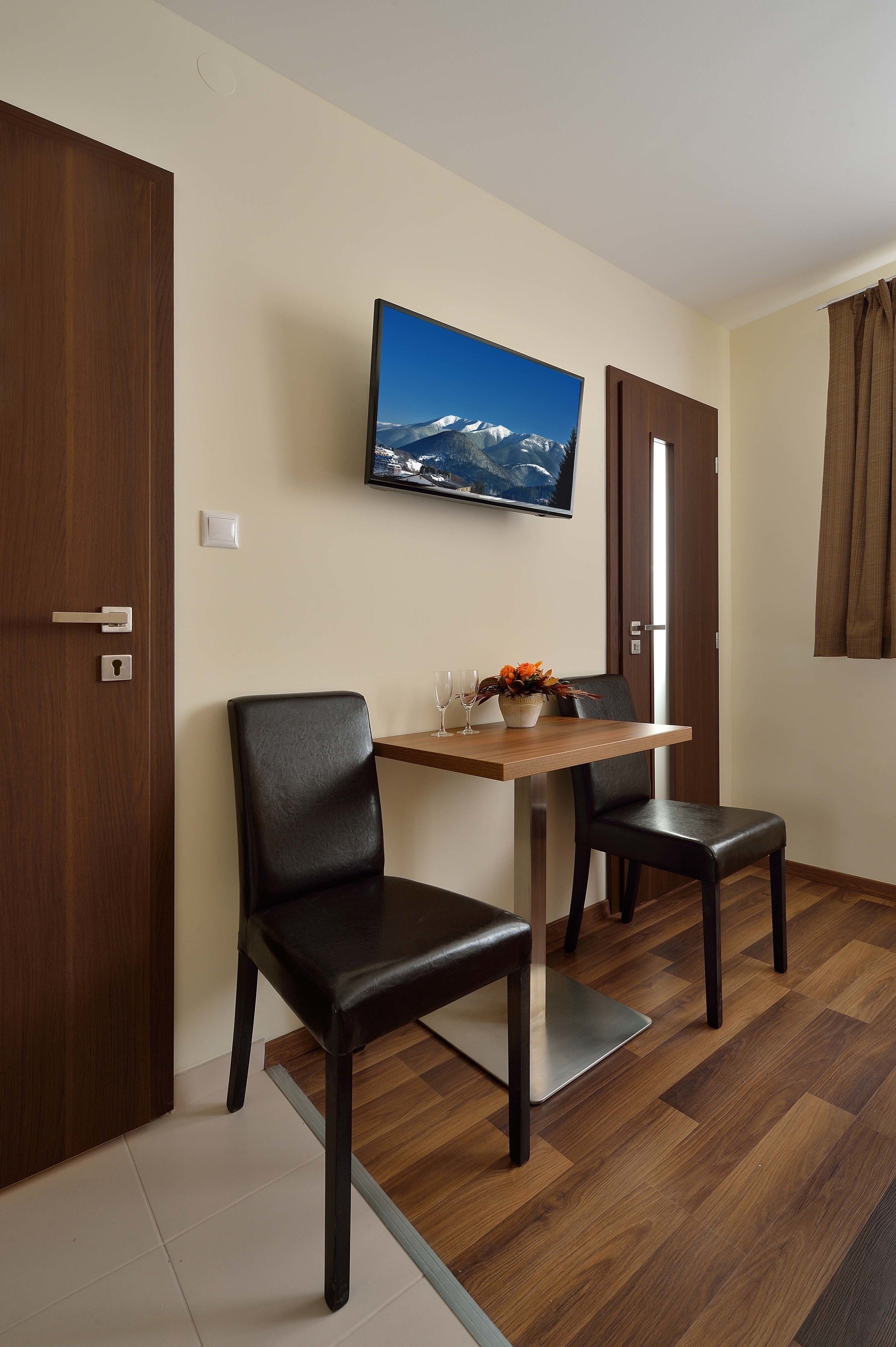 Een kamer met een houten eettafel, twee zwarte stoelen, een aan de muur bevestigde televisie en een deur met een handgreep.