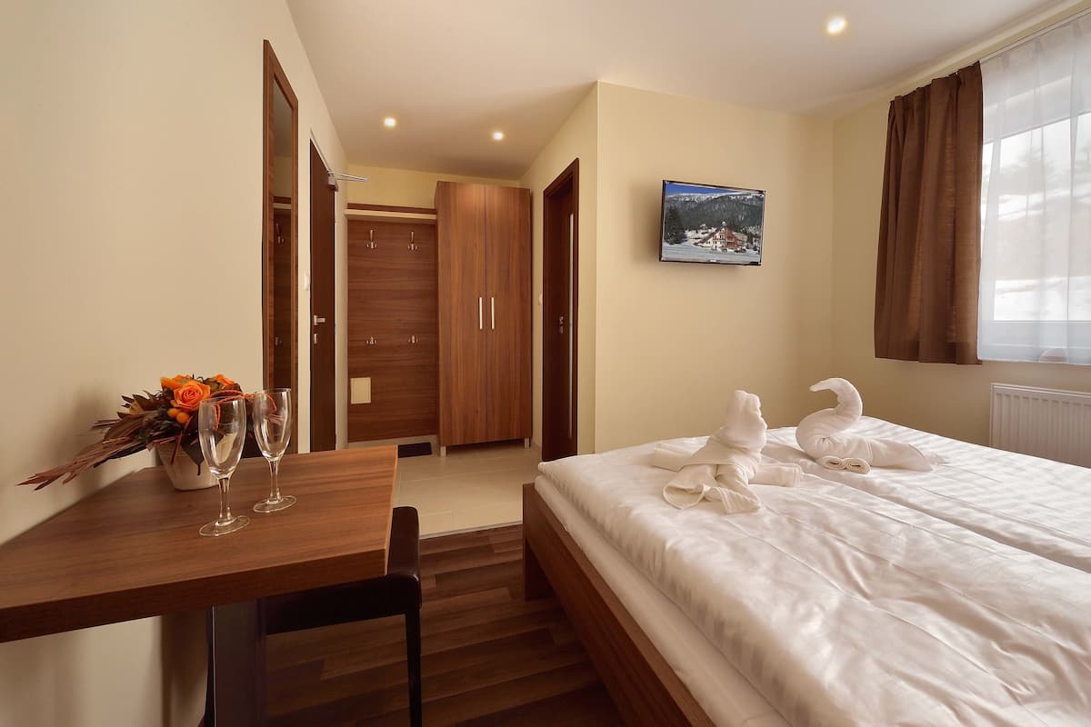 A hotel room with a wooden desk, two wine glasses, a bouquet of flowers, a bed with white linens, and a television on the wall.