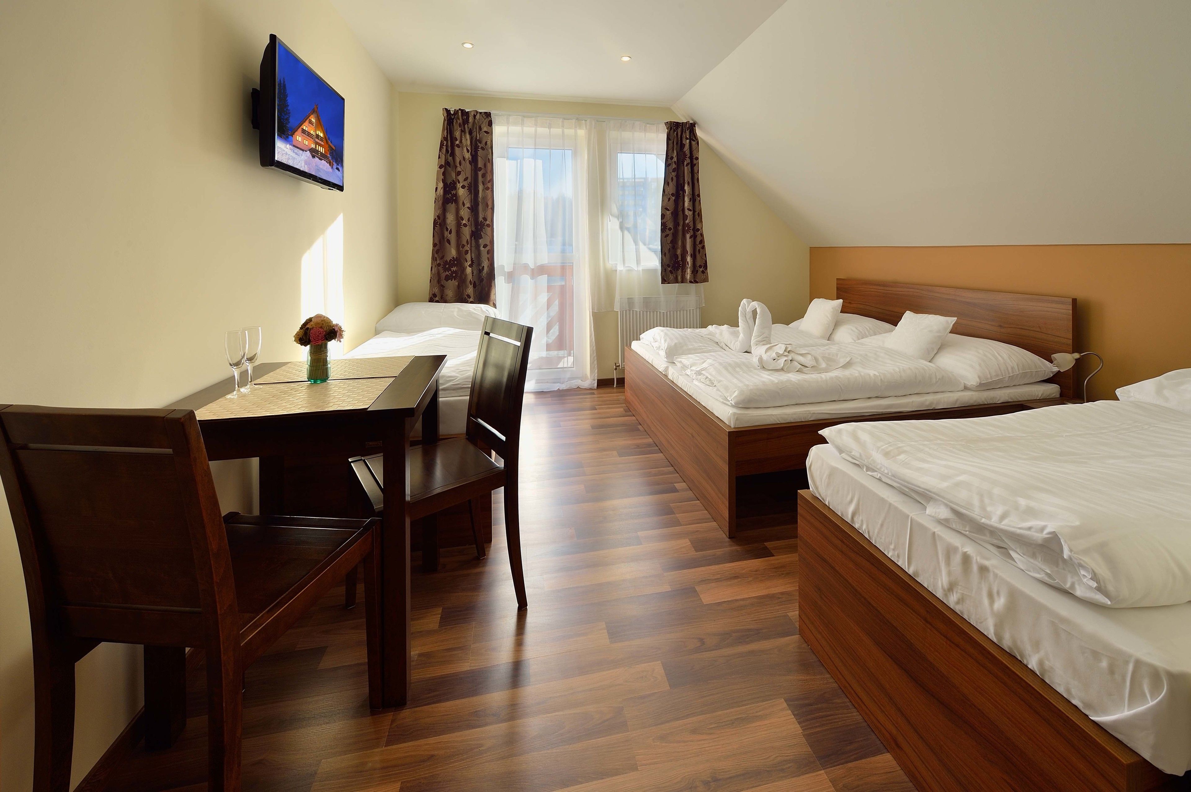 Habitación de hotel con dos camas individuales, un escritorio de madera con sillas, un televisor montado en la pared y una ventana con cortinas.