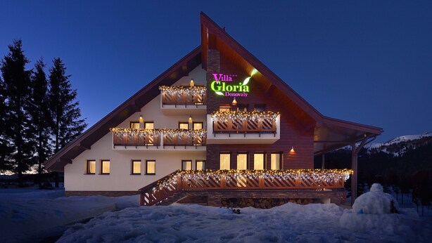 Un edificio de dos pisos con un letrero que dice "Villa Gloria Donoval". Tiene fachada de madera, balcones con barandillas y el suelo cubierto de nieve.