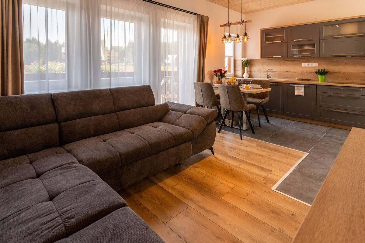 A modern living room with a sectional sofa, a dining area with chairs, a kitchen with grey cabinets, and a large window with sheer curtains.