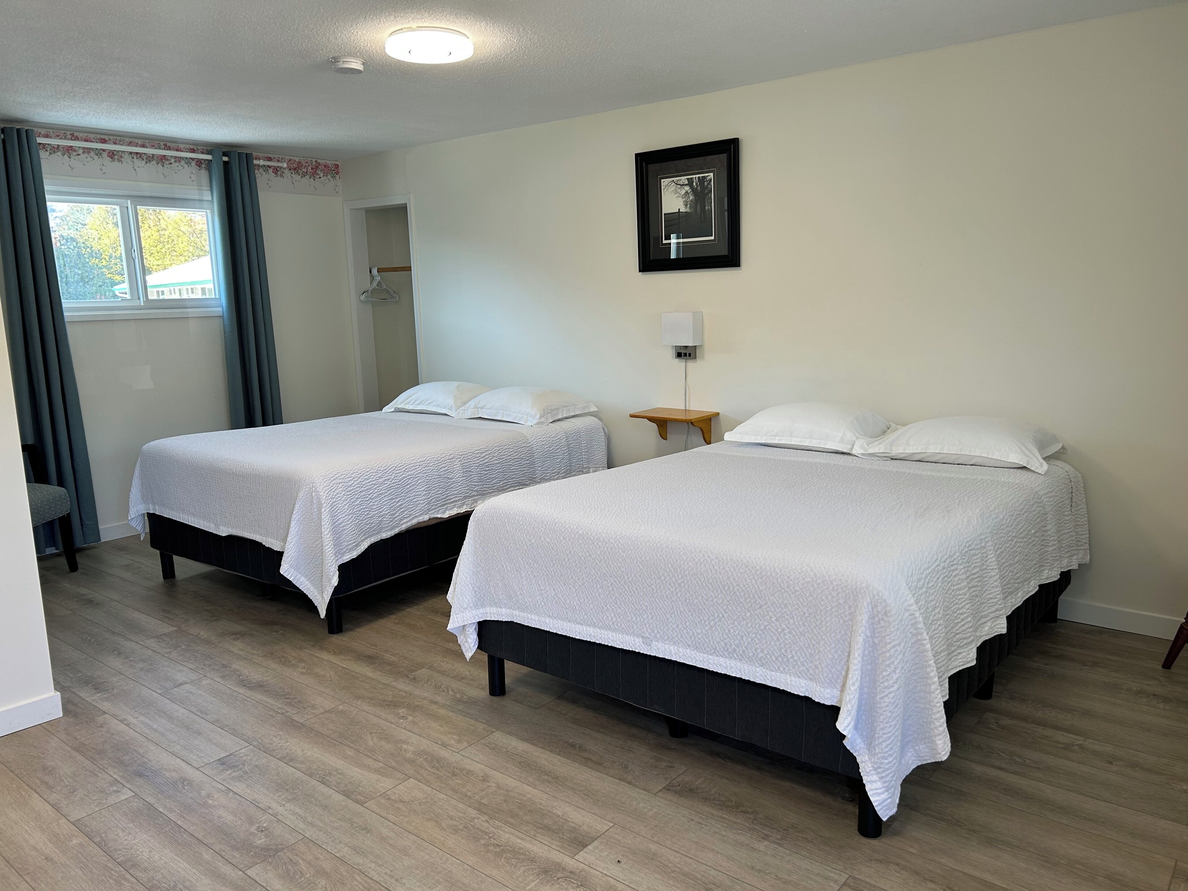 Ett hotellrum med två sängar, trägolv, ett fönster med gardiner, en inramad tavla på väggen och ett nattduksbord med en lampa.
