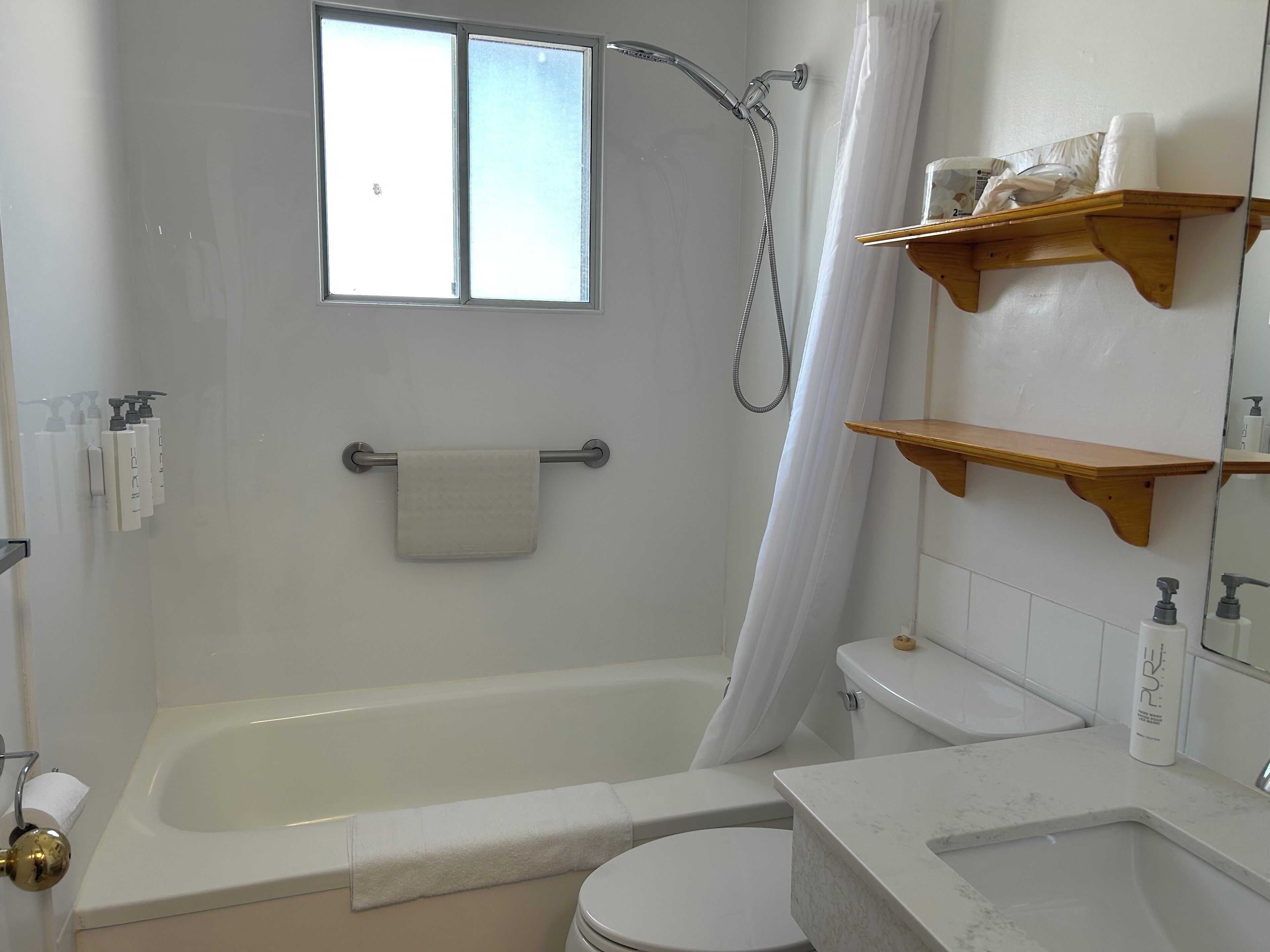 A bathroom with a bathtub, toilet, and a shower. There are wooden shelves above the toilet and a window above the bathtub.