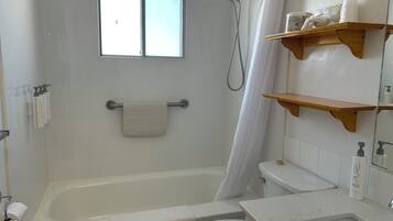 A bathroom with a bathtub, toilet, and a shower. There are wooden shelves above the toilet and a window above the bathtub.