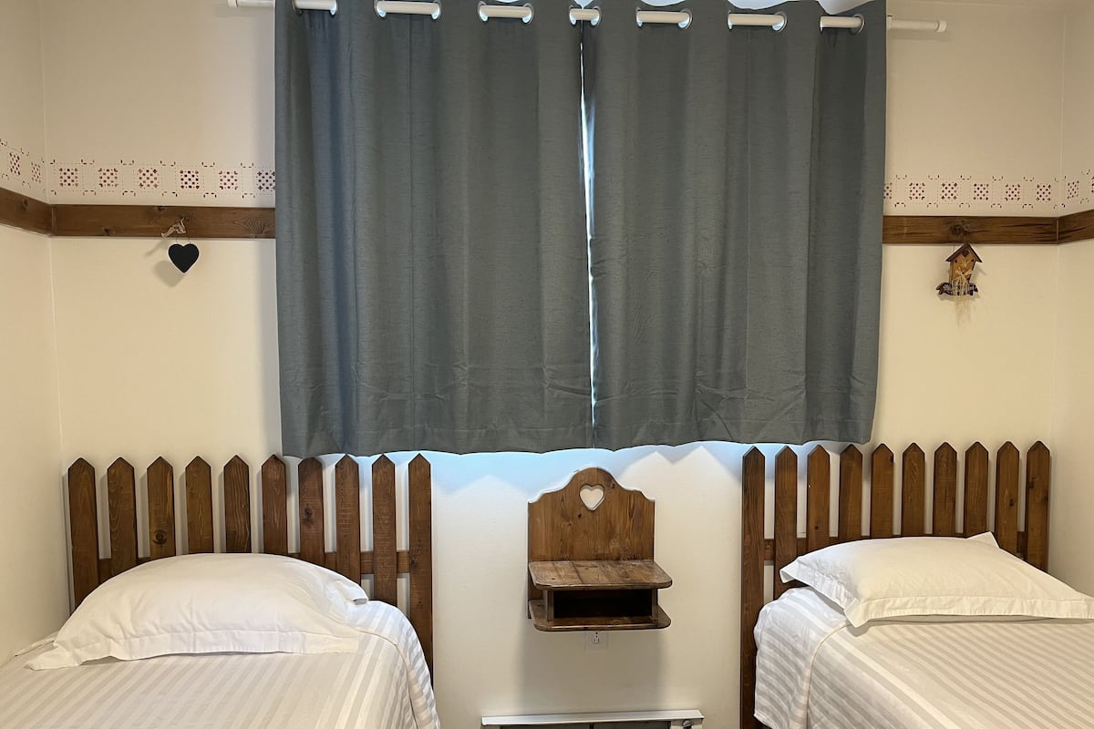 Una habitación con dos camas, cabecero de madera, un estante pequeño y una ventana con cortinas.