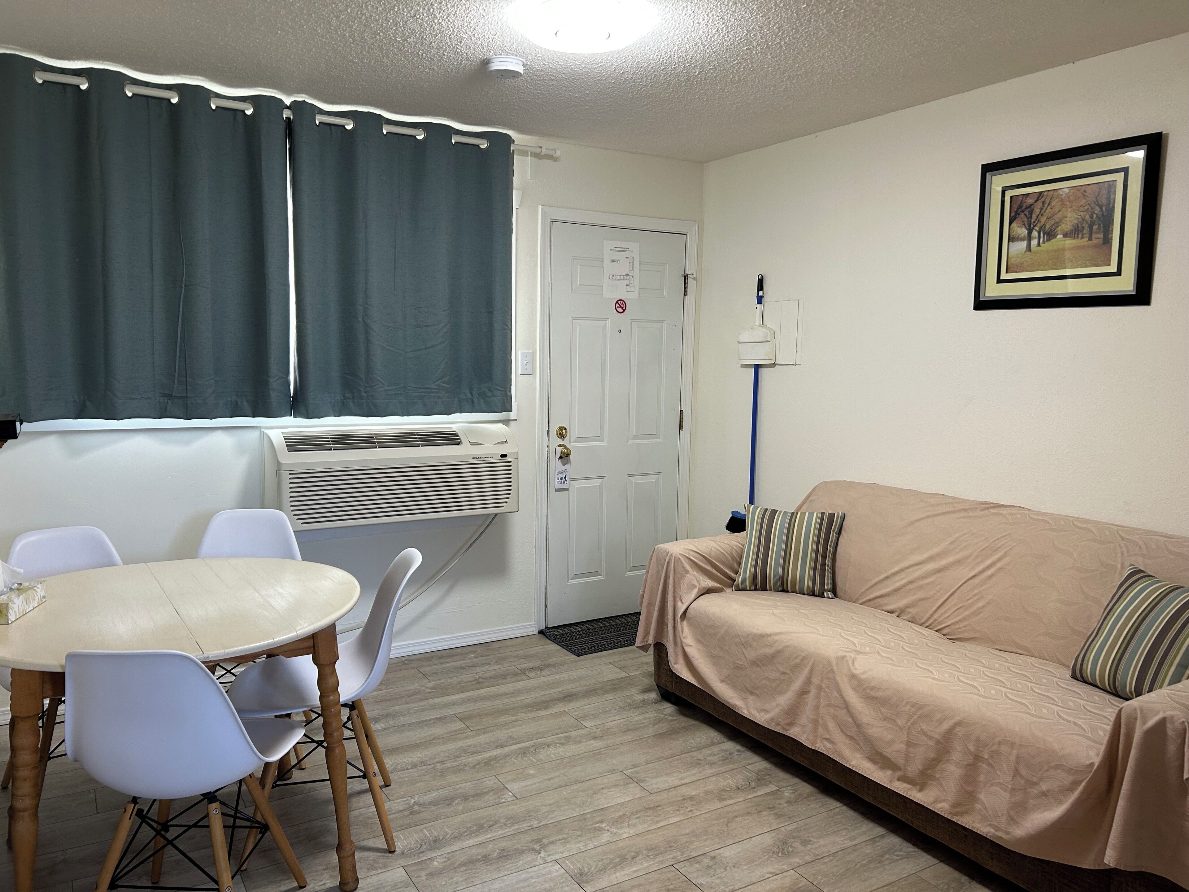 A small, furnished room with a dining table, chairs, a sofa, a window with curtains, an air conditioning unit, and a door with a lock.