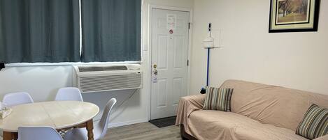 A small, furnished room with a dining table, chairs, a sofa, a window with curtains, an air conditioning unit, and a door with a lock.